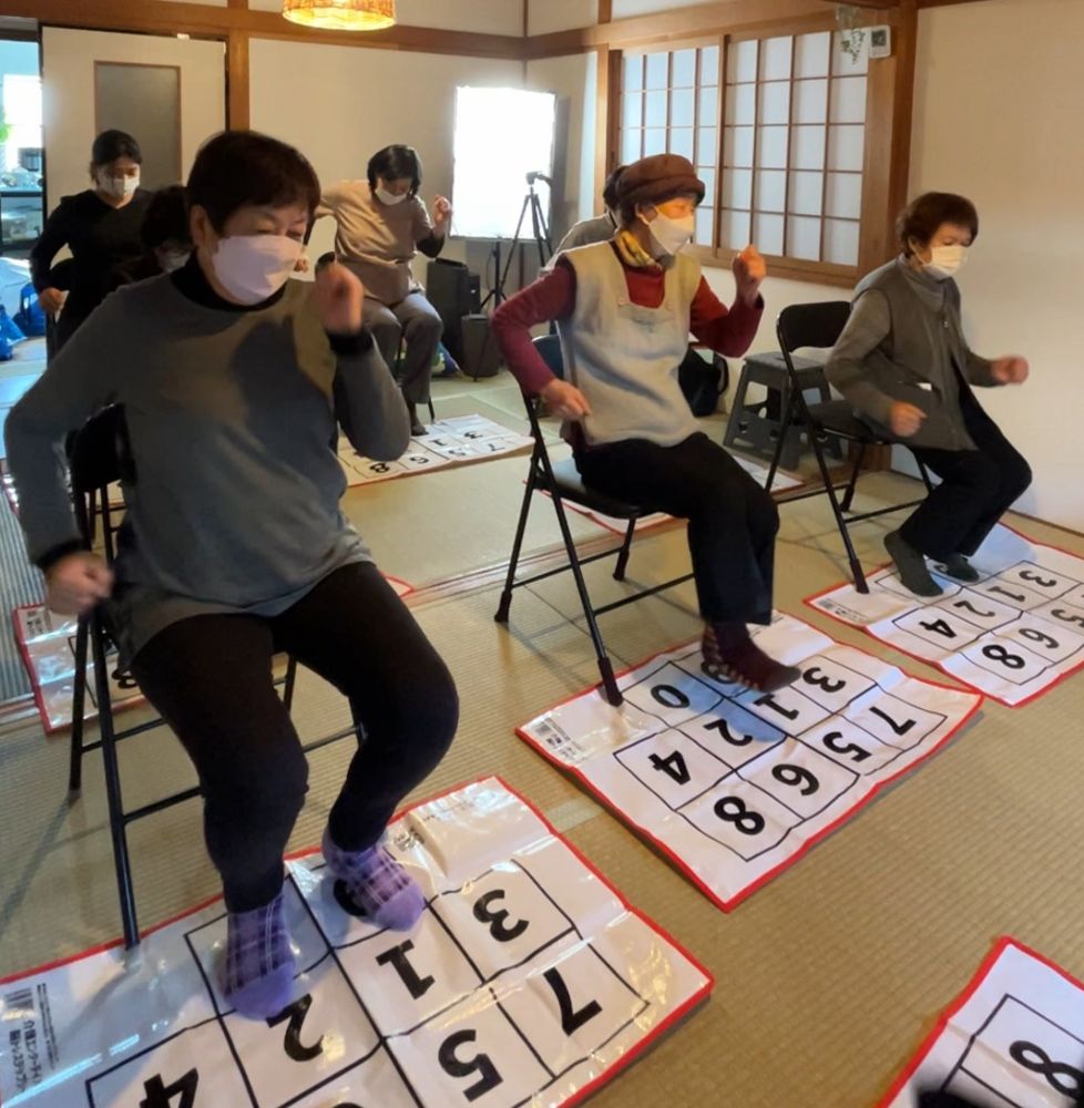 和室の体操教室で、椅子に座ったまま足元の脳トレステップシートを踏む高齢者の皆さん