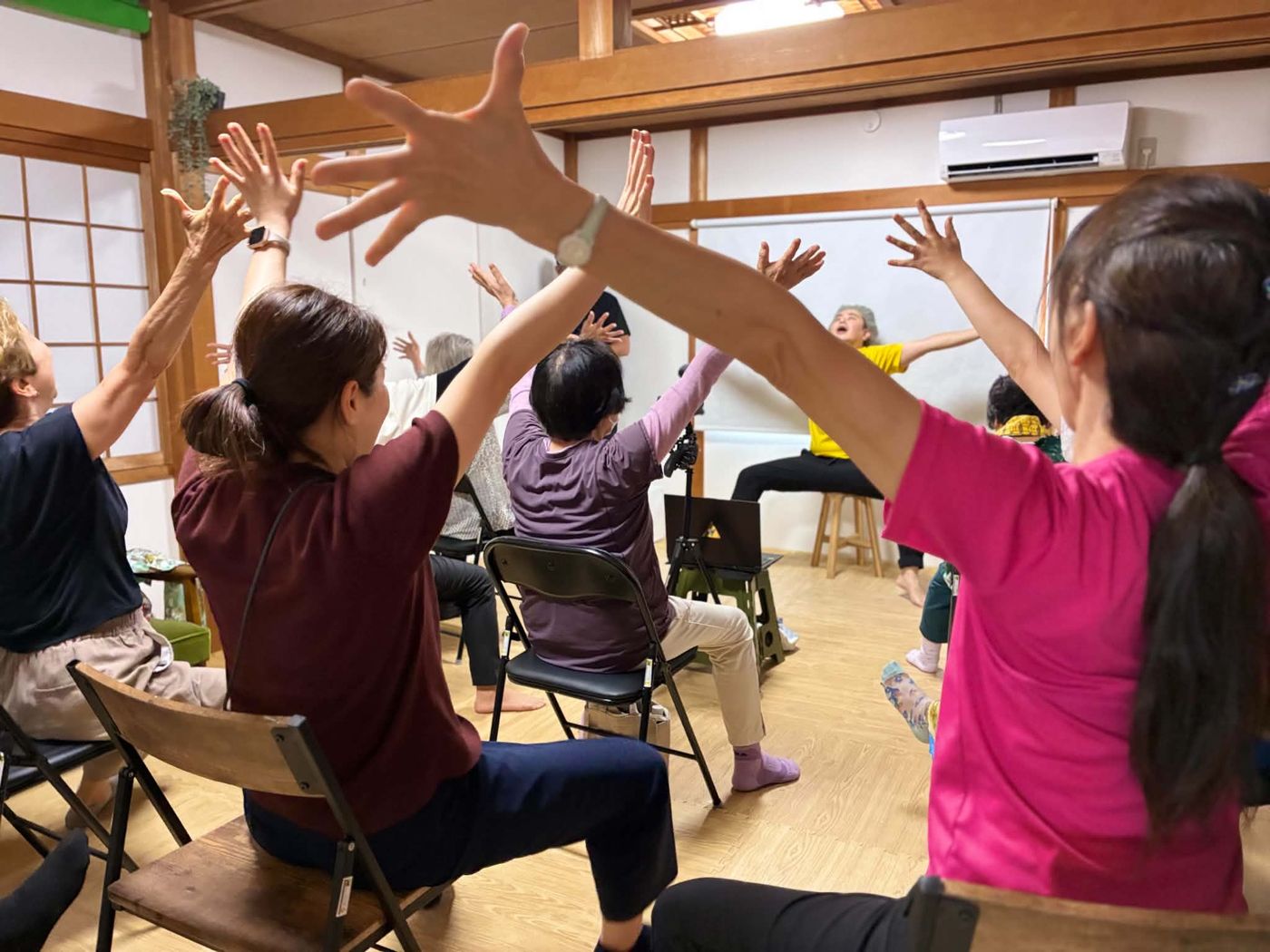 おふとんハウスの室内で、椅子に座った参加者が両手を上げて体操している様子