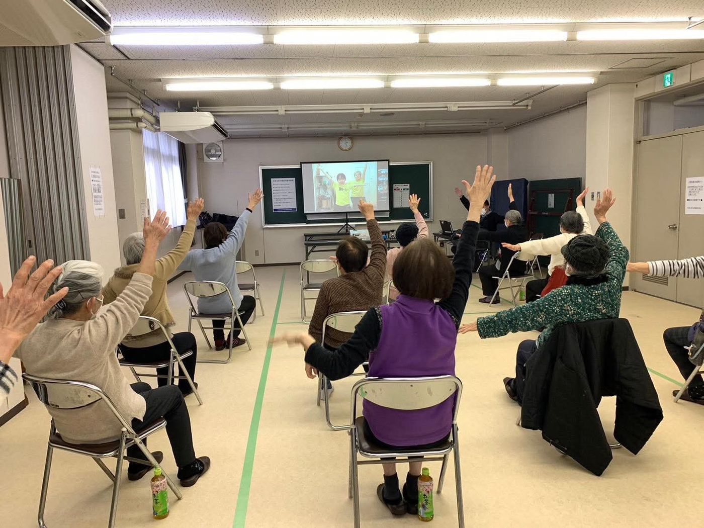 公民館の一室で、参加者がスクリーンに映った体操に合わせて手を上げている様子