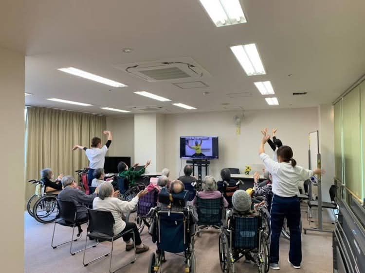 施設のフロアで、画面に映った体操に合わせて車椅子の方も含めて全員が手を上げている様子