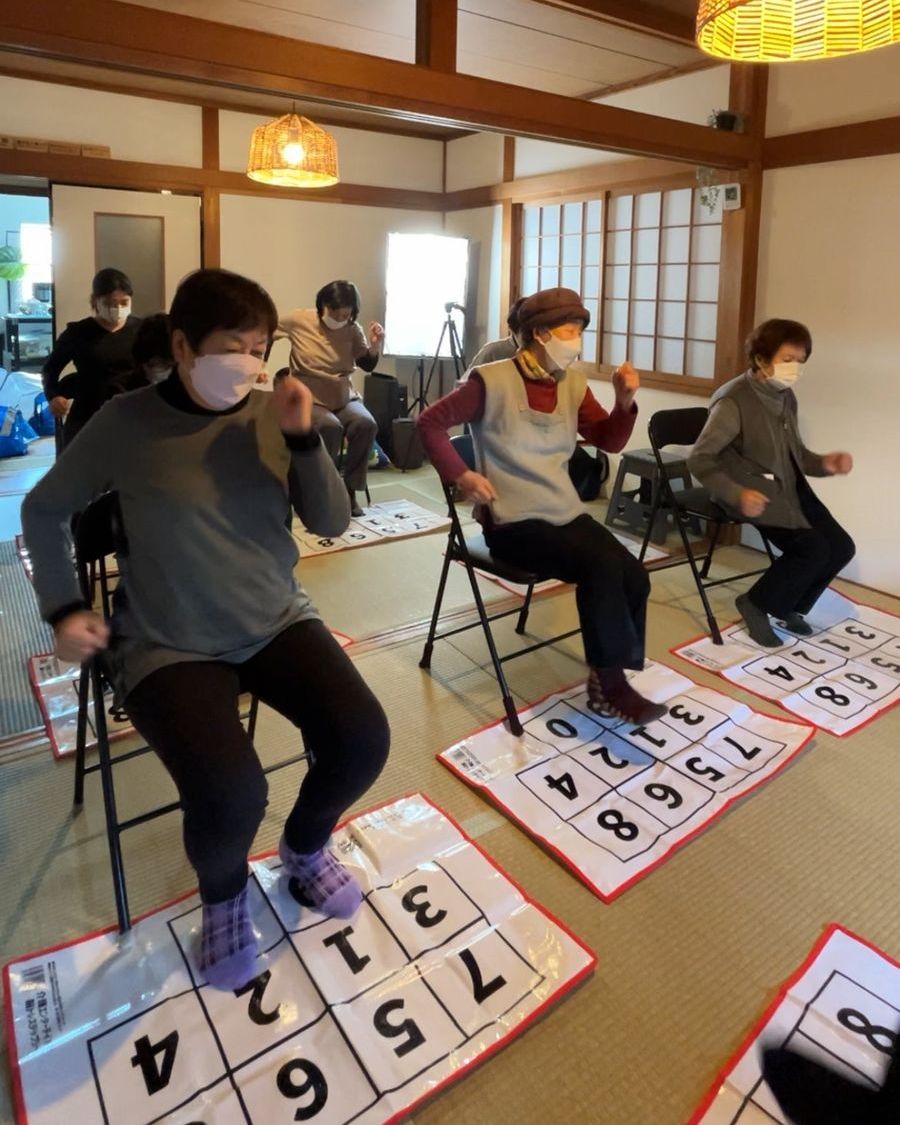 和室で椅子に座り、足元の脳トレステップシートを踏みながら体操する高齢者の皆さん